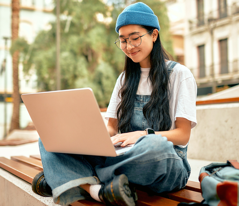Female student sitting outside with her laptop on a bench typing.
